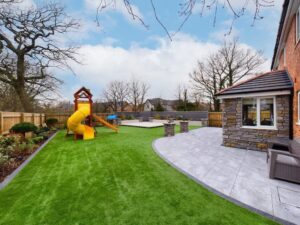 A landscaped backyard featuring artificial grass, a children's playset with a slide, a curved grey tiled patio, and a stone-clad house extension.