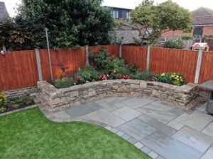 A landscaped garden featuring a curved stone retaining wall with colorful flowers, a paved patio, artificial grass, and wooden fencing.