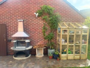 Brick wall and cobblestone patio featuring a greenhouse and BBQ, with a green potted plant placed beside the BBQ
