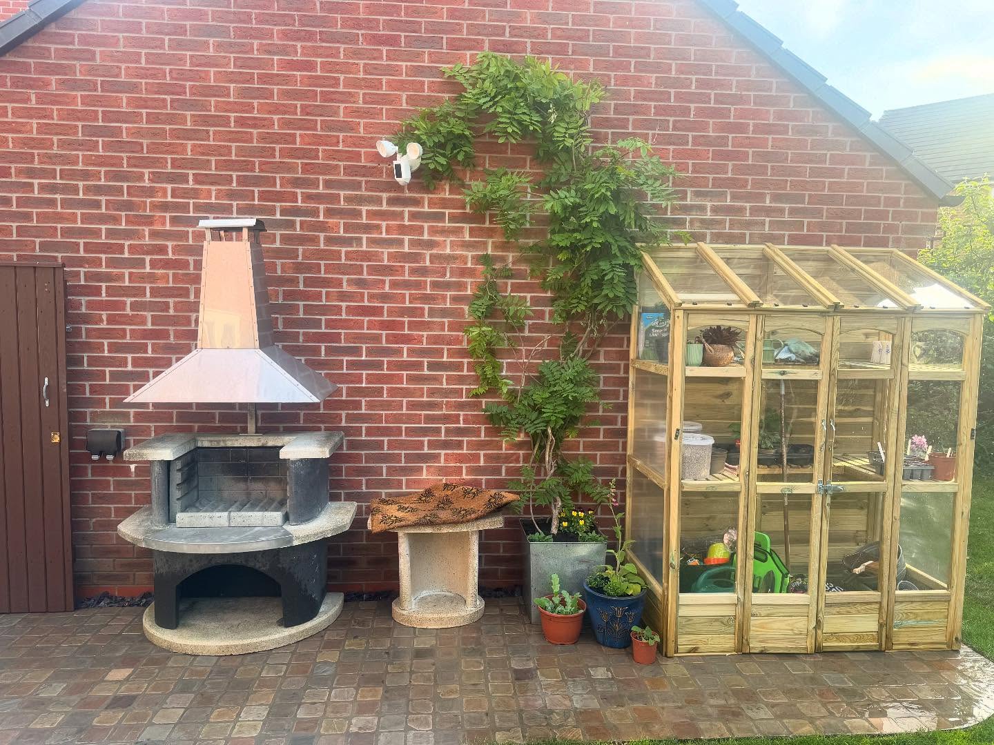 Brick wall and cobblestone patio featuring a greenhouse and BBQ, with a green potted plant placed beside the BBQ